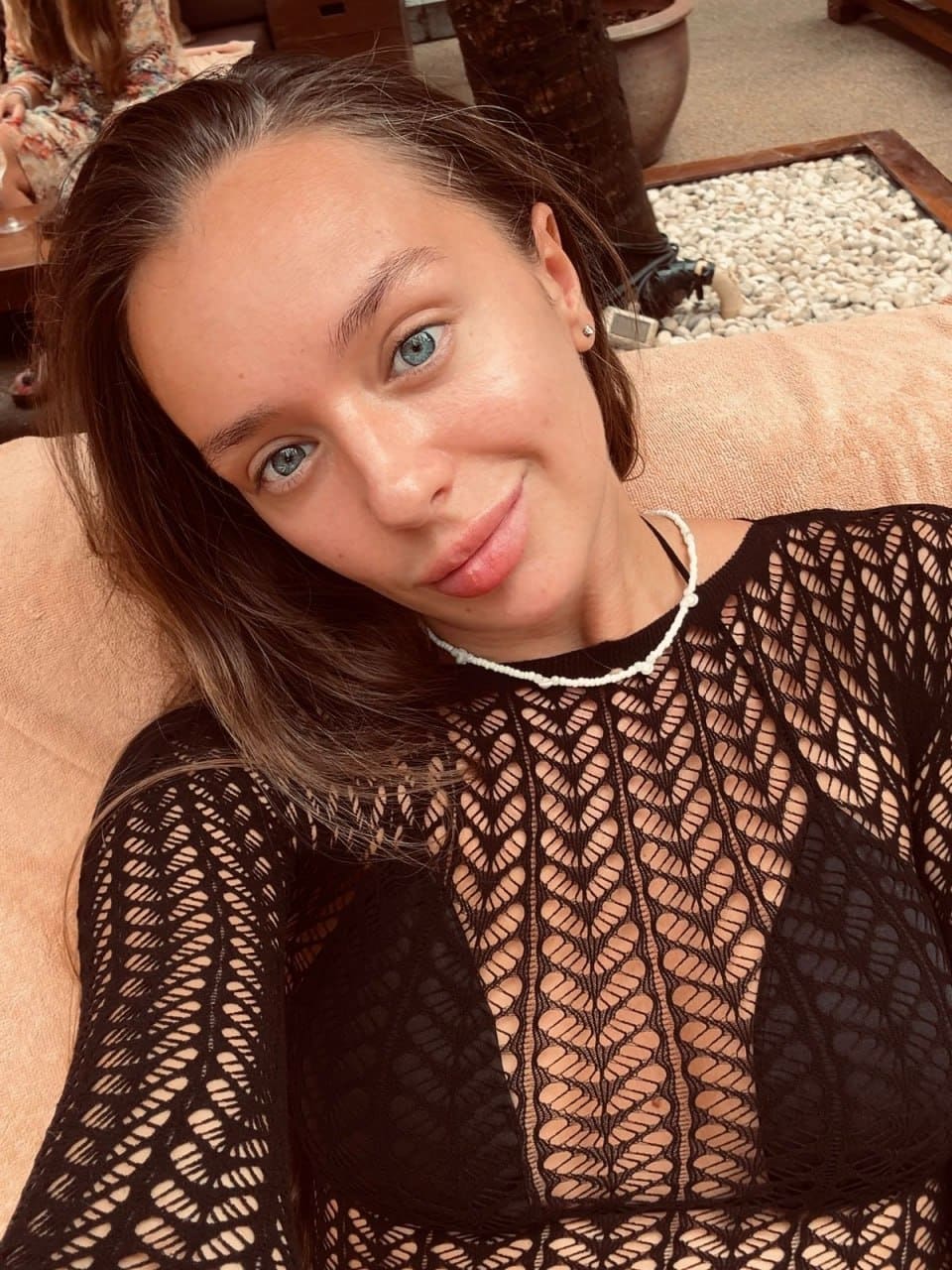 Close-up selfie of woman with pulled-back hair in black mesh top and bra, reclining on a peach-colored sofa with beaded necklace.