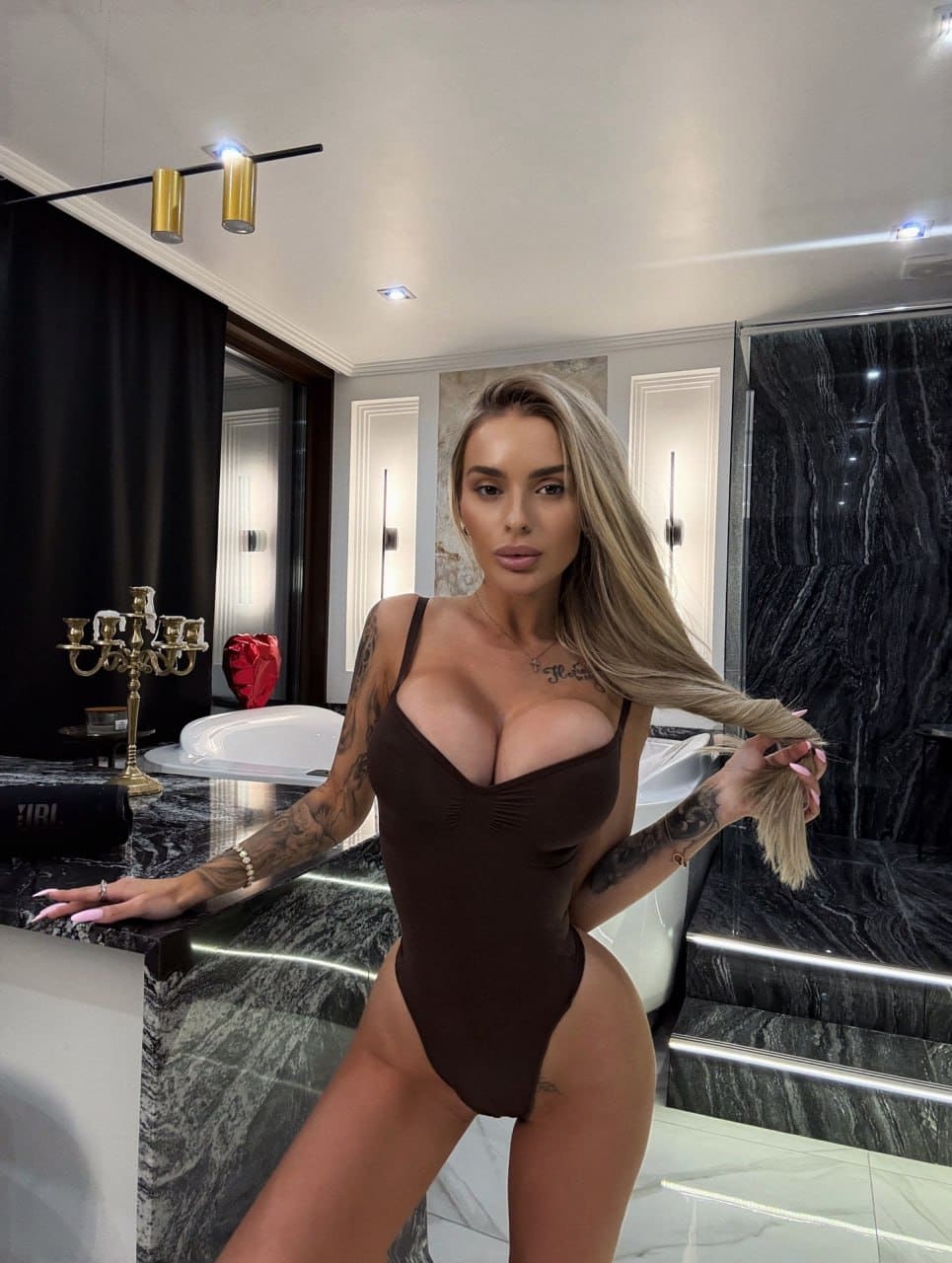 A woman in a dark brown plunging bodysuit poses in a marble bathroom, resting a hand on the counter.