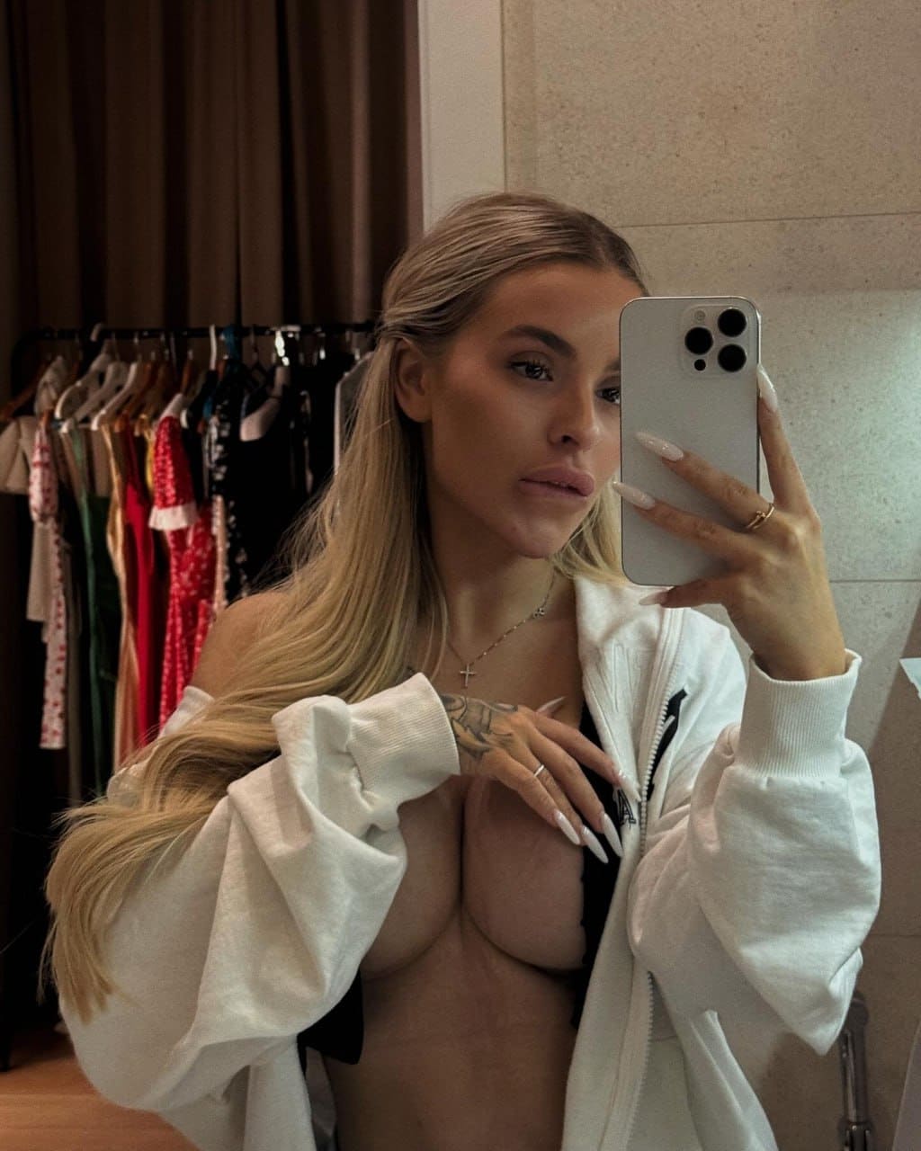 Mirror selfie of a woman in an open white hoodie and black thong, standing before a clothing rack.