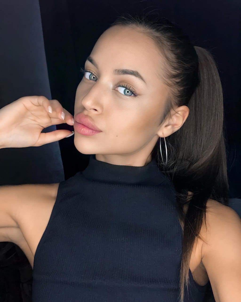 A woman with a high ponytail wears a black turtleneck and silver hoops, raising one hand near her face against a dark background.