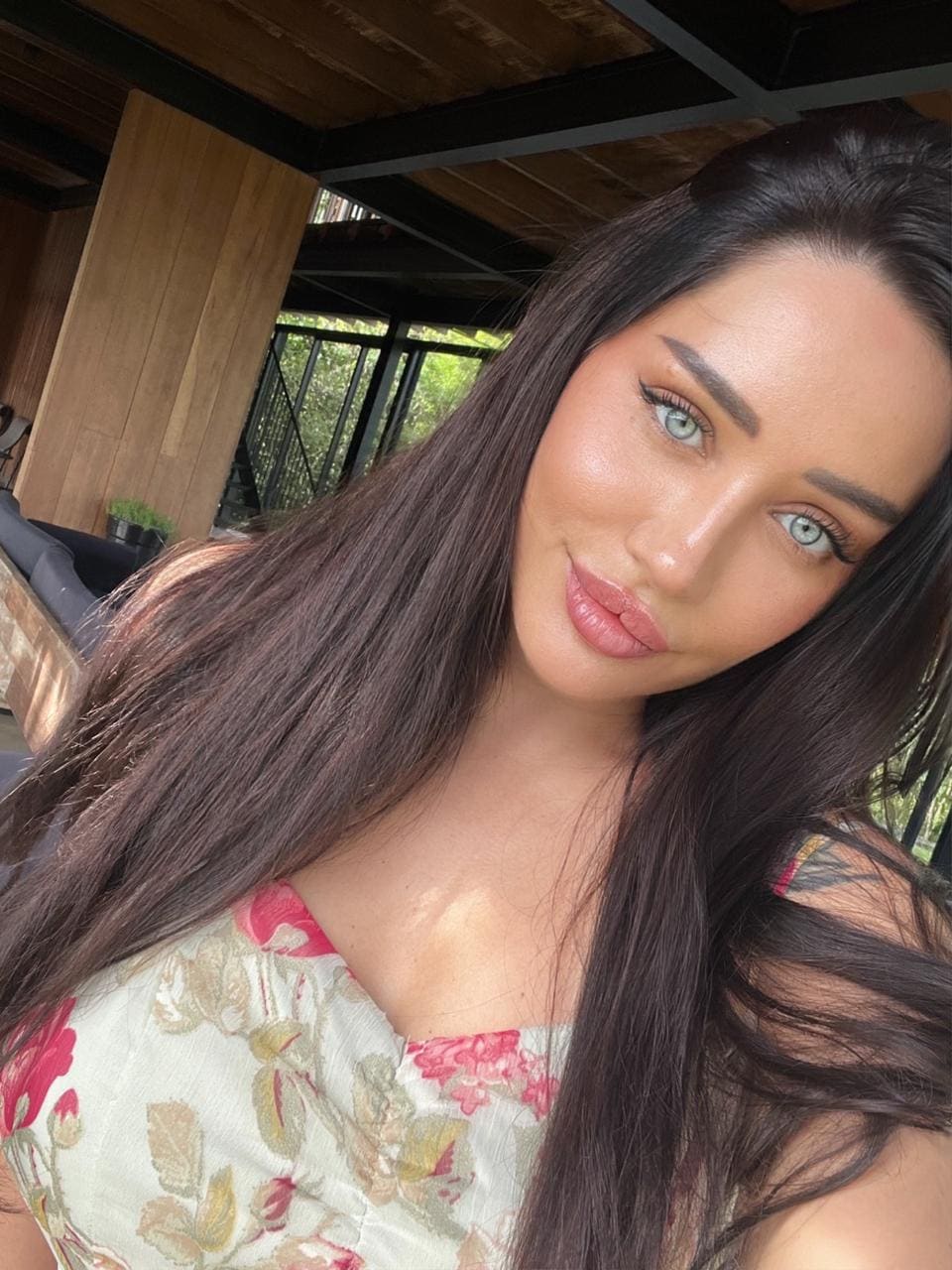 A close-up selfie shows a woman in a floral sweetheart-neckline top against wooden paneling and greenery, exposing shoulders.