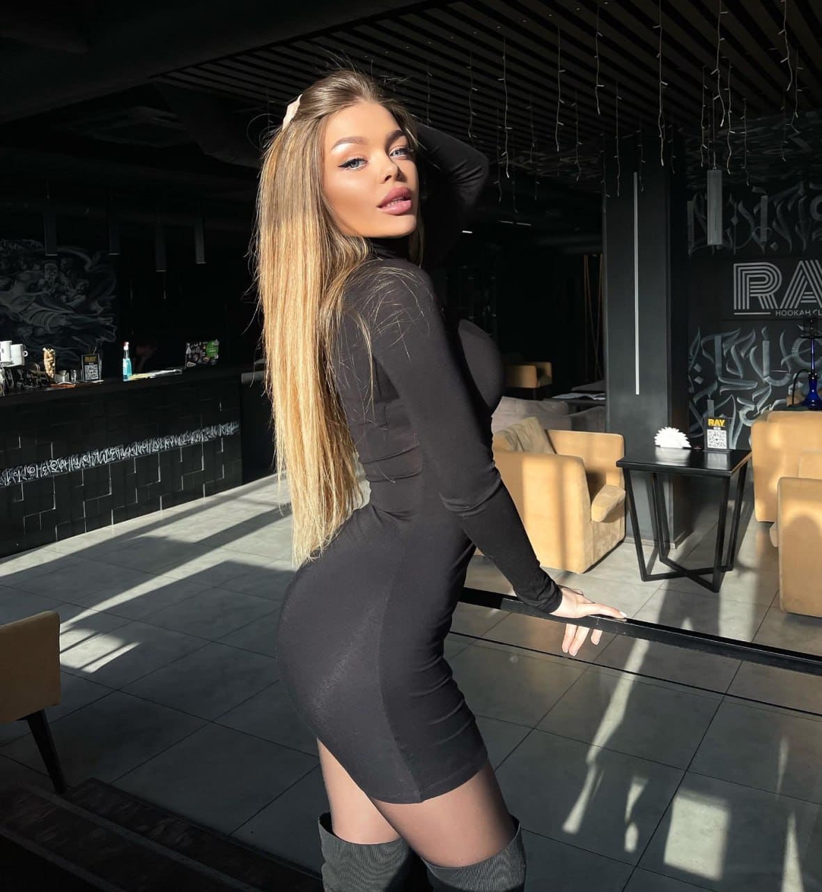 Woman in a black mini dress and thigh-high boots stands in profile in a lounge, one hand on her head, the other on a surface.