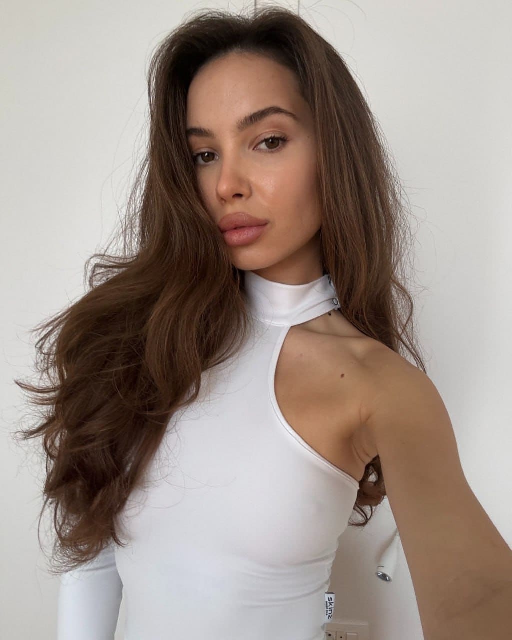 Lolina stands against a plain light wall, wearing a white high-neck sleeveless top with a shoulder cutout and long wavy brown hair.