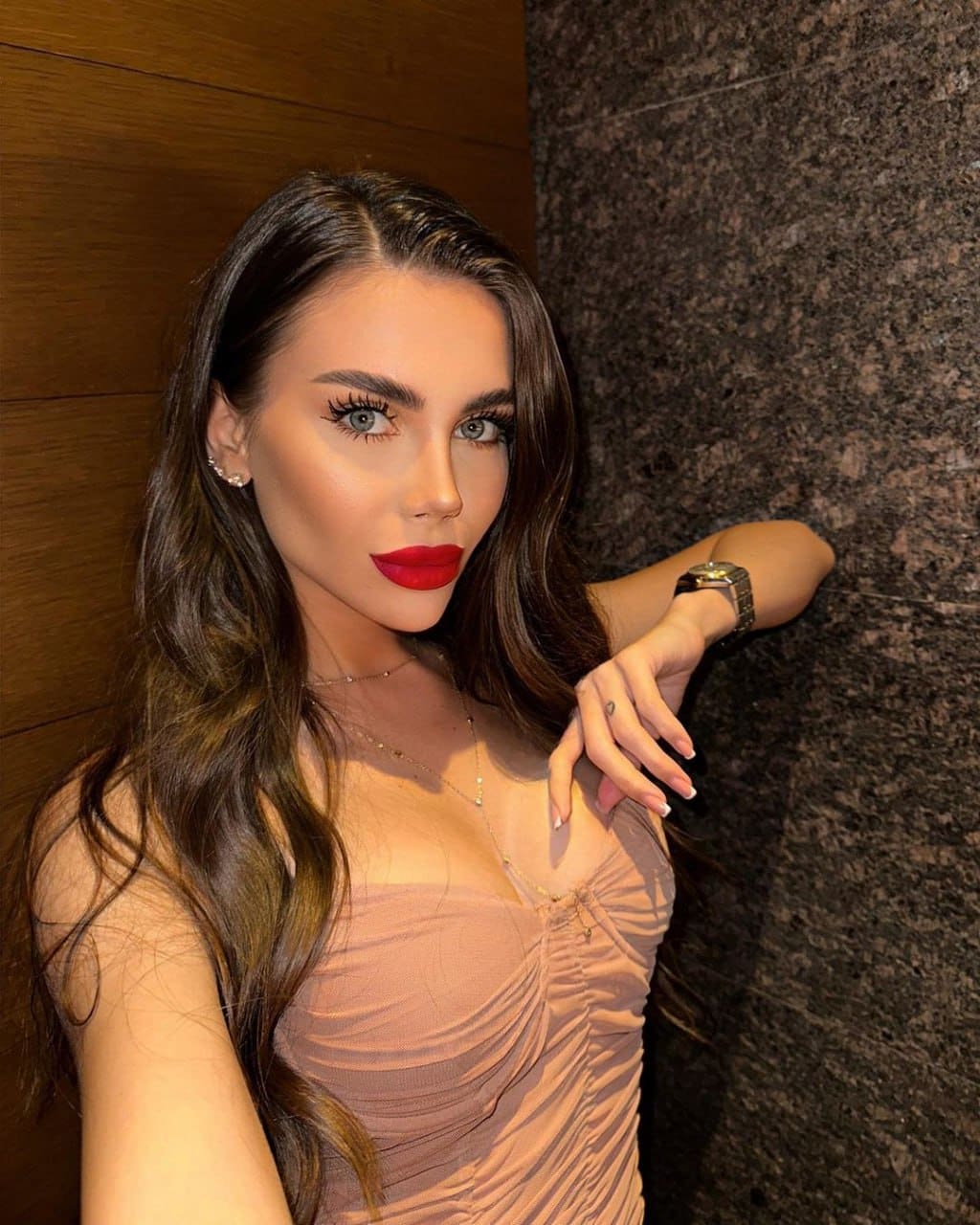 A woman with dark wavy hair and red lipstick wears a strapless beige top, posing against wood paneling and dark stone with a watch visible.