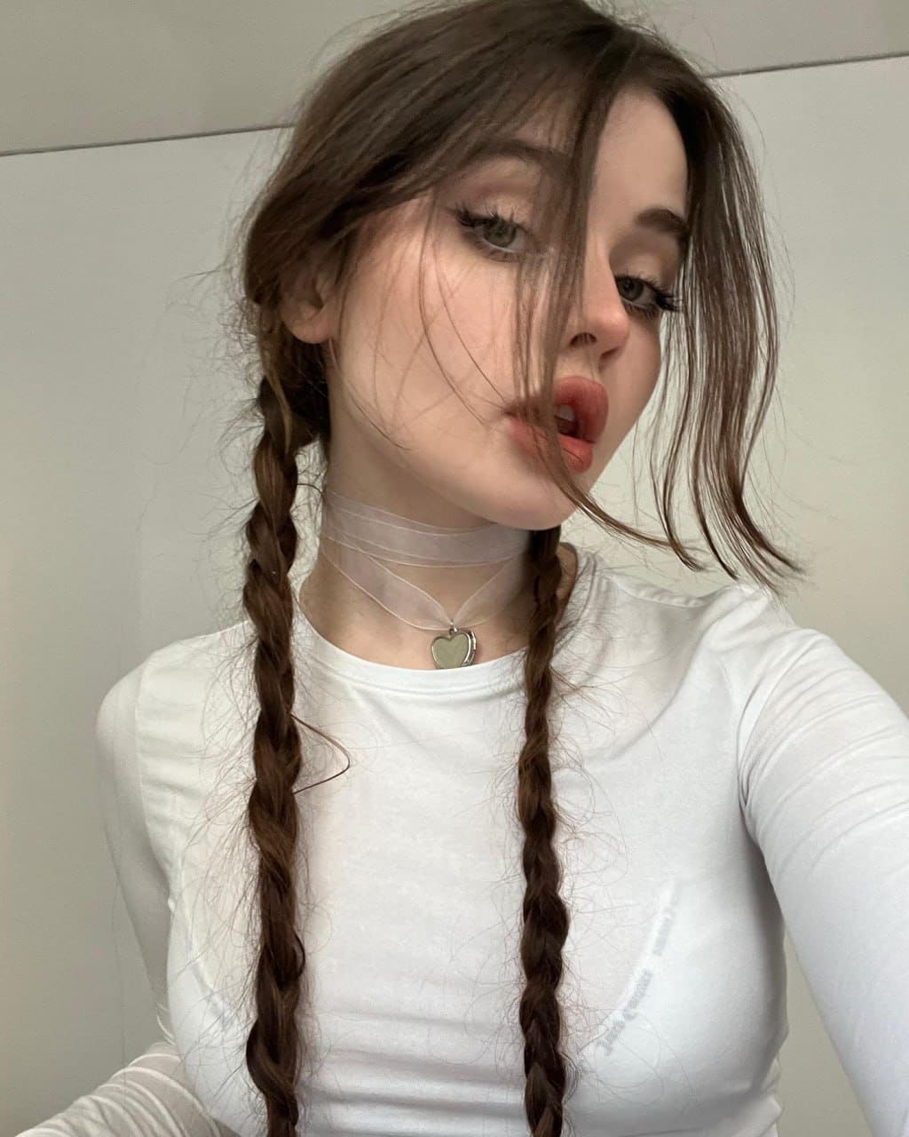 Valentina with long brown hair in two braids wears a white long-sleeved top and heart choker, posing for a selfie against a white background.