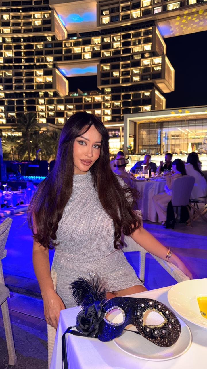 At an outdoor table at night, a woman in a short silver sequined dress sits with a black masquerade mask resting on the surface.