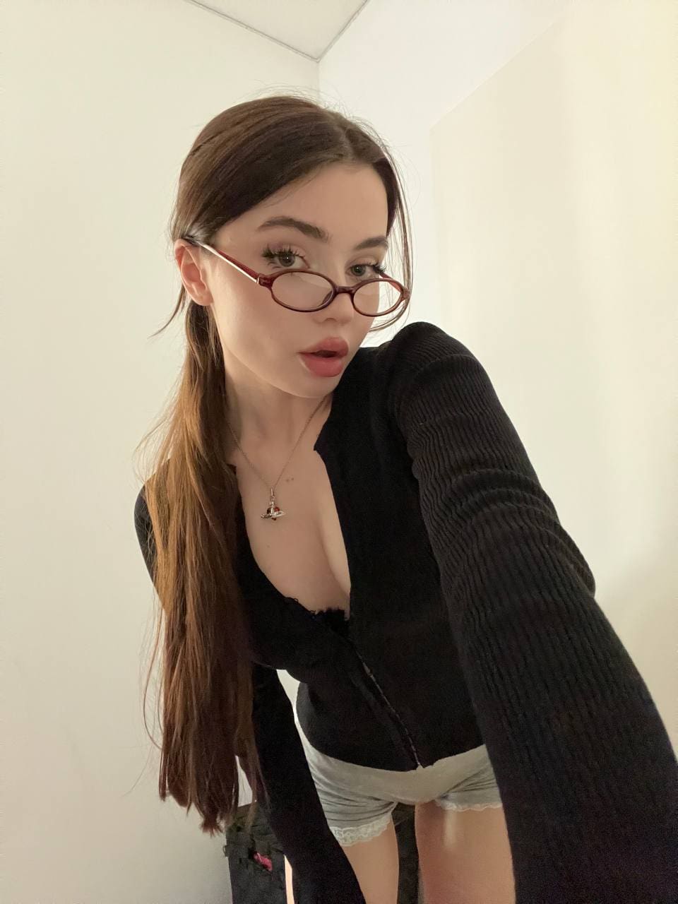 A woman with long brown hair and oval glasses leans forward, wearing a black long-sleeved top with a deep neckline and grey lace-trimmed shorts.