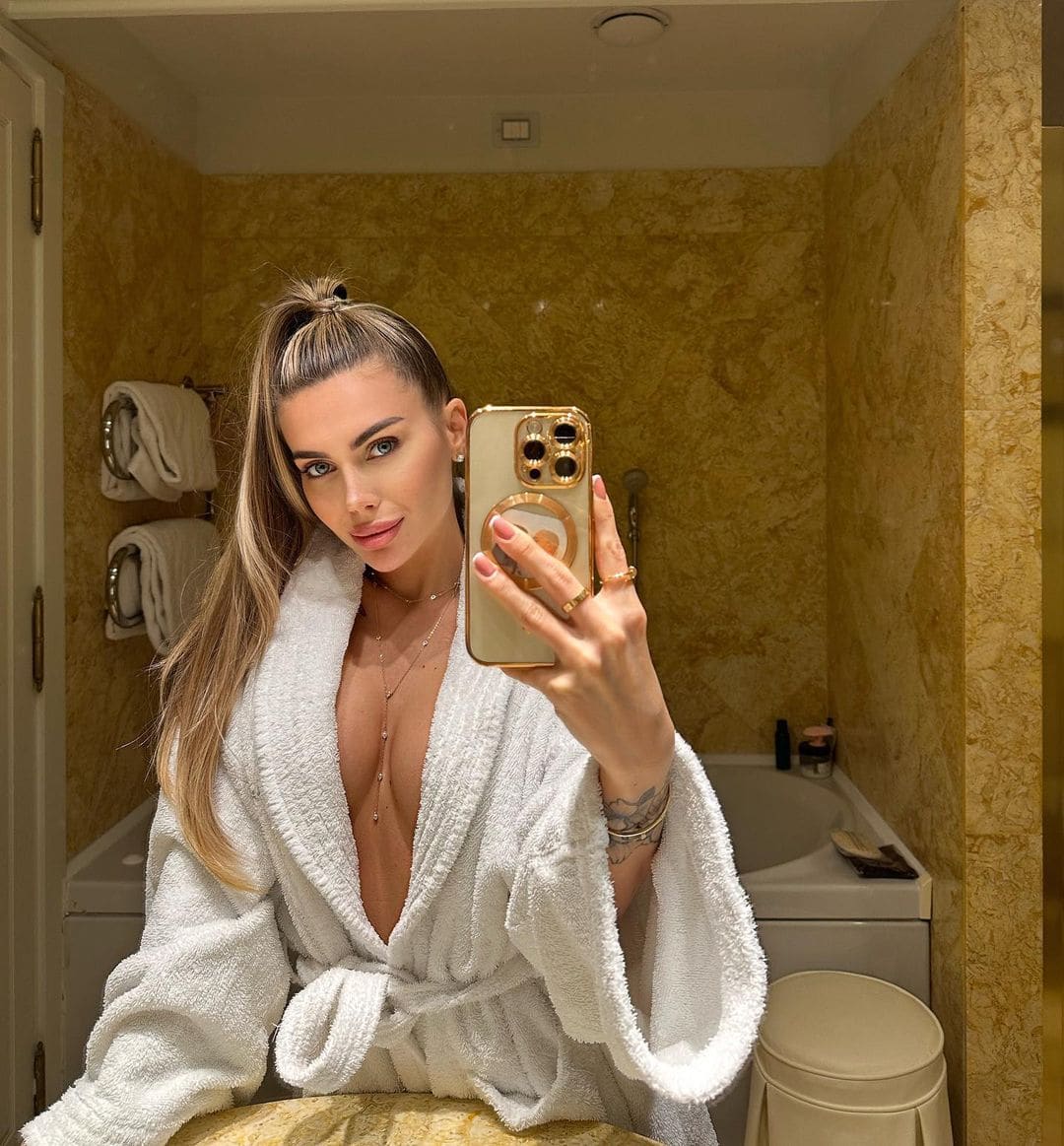 A blonde woman with a high ponytail takes a bathroom mirror selfie wearing an open white terry cloth bathrobe and a long body chain.