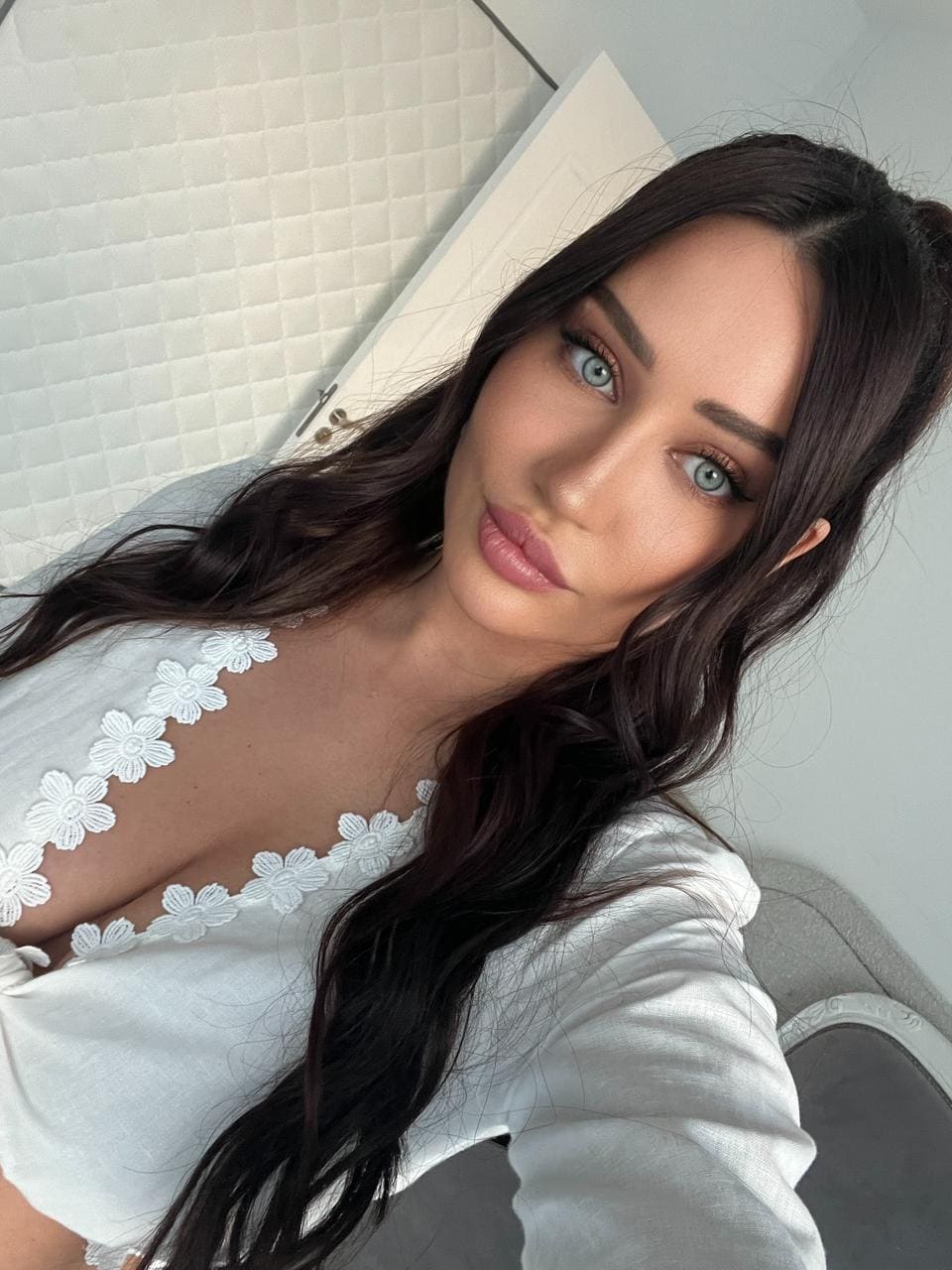 A selfie features a woman in a white top with a deep plunging neckline and floral lace trim, against a white quilted wall and door.