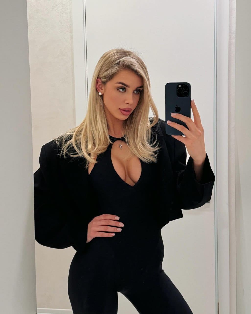 A woman in a black blazer and keyhole bodysuit poses with one hand on her hip for a mirror selfie.