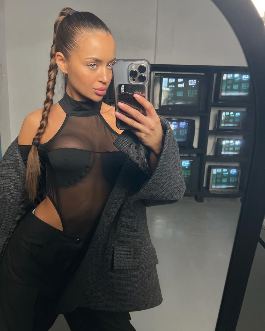 A woman with a braided ponytail takes a mirror selfie in a sheer black bodysuit, trousers, and a grey blazer, standing before a rack of vintage monitors.