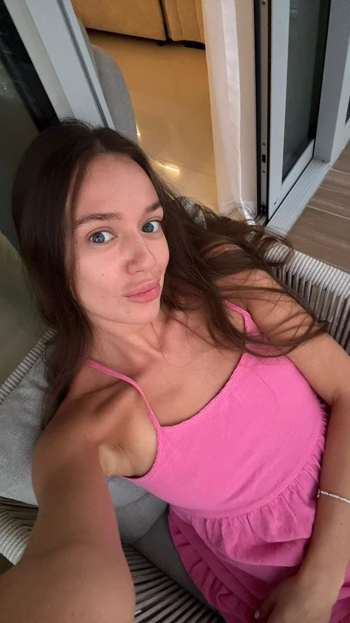 Woman with long dark hair reclining on wicker sofa, wearing a pink ruffled spaghetti-strap top and looking directly at the camera.