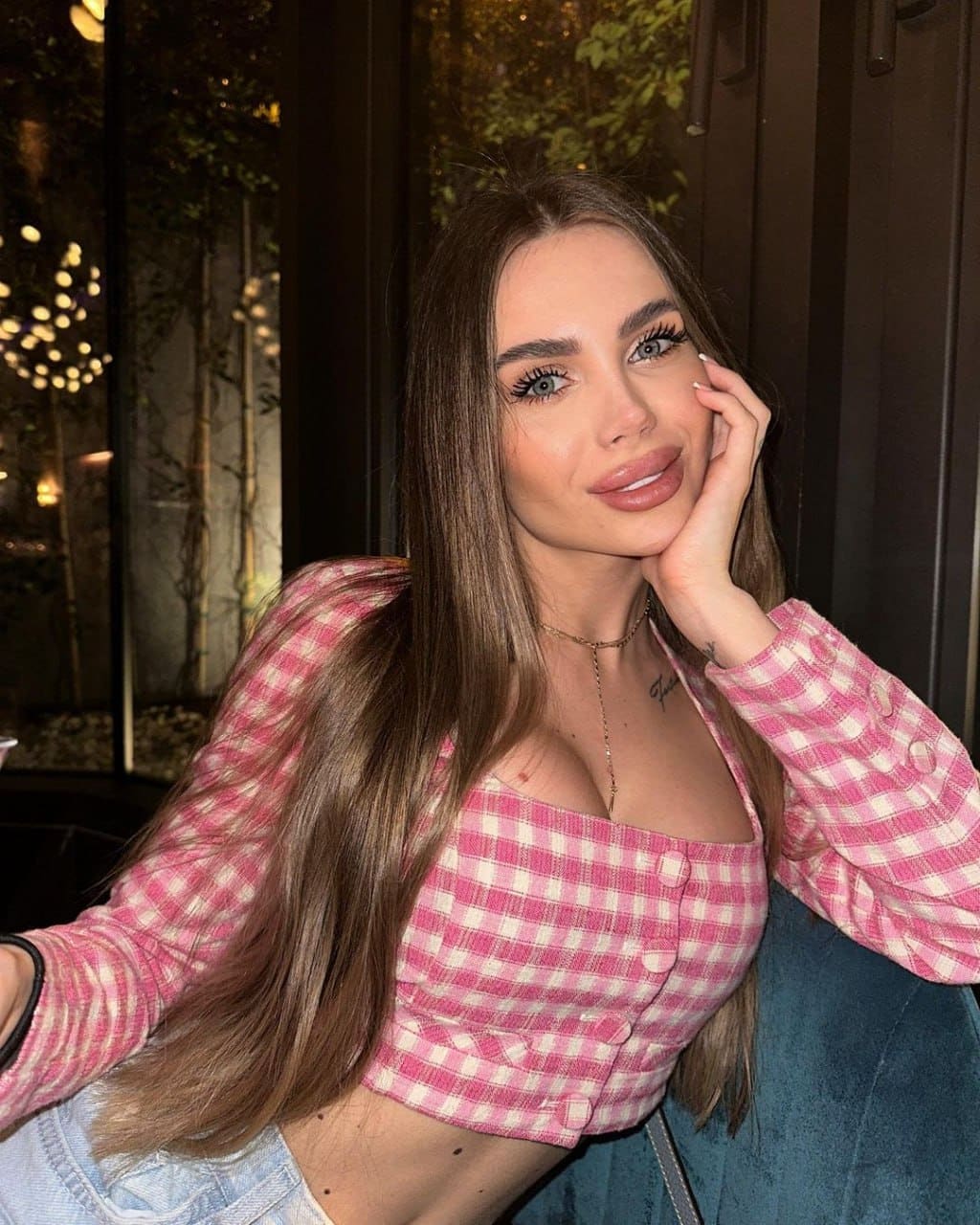 Lyra with long brown hair rests a hand on her cheek, wearing a pink gingham crop top and denim against a dark background with blurred lights.